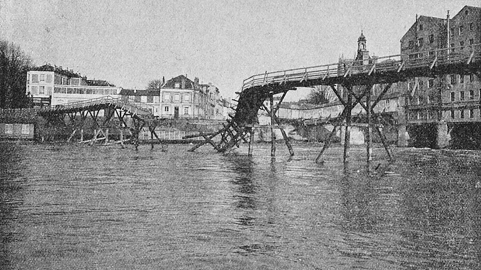Zniszczona drewniana kładka nad rzeką, za nią kamienice miasteczka.
